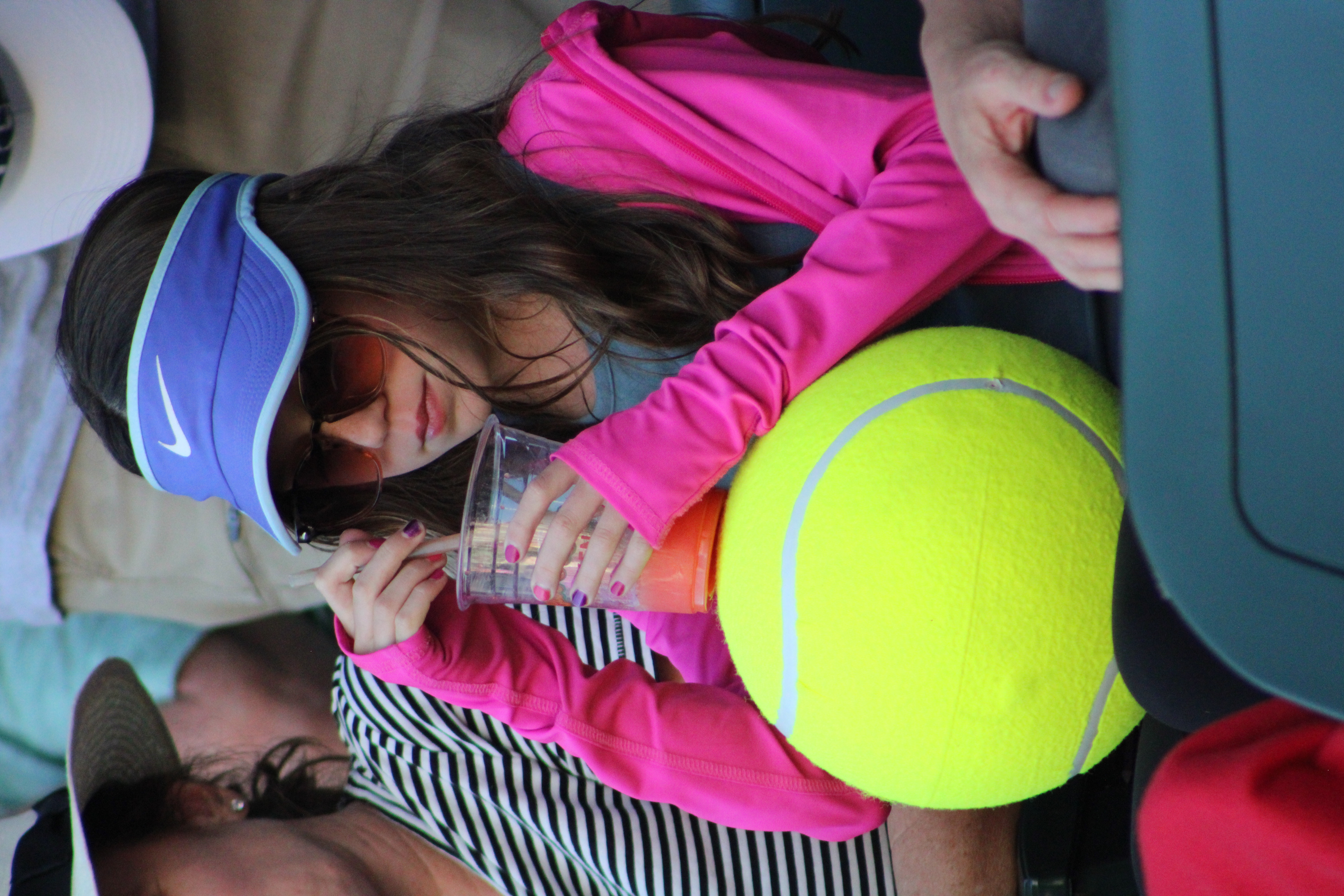 Young girl spectator with her oversized tennis ball and stirring her beverage.